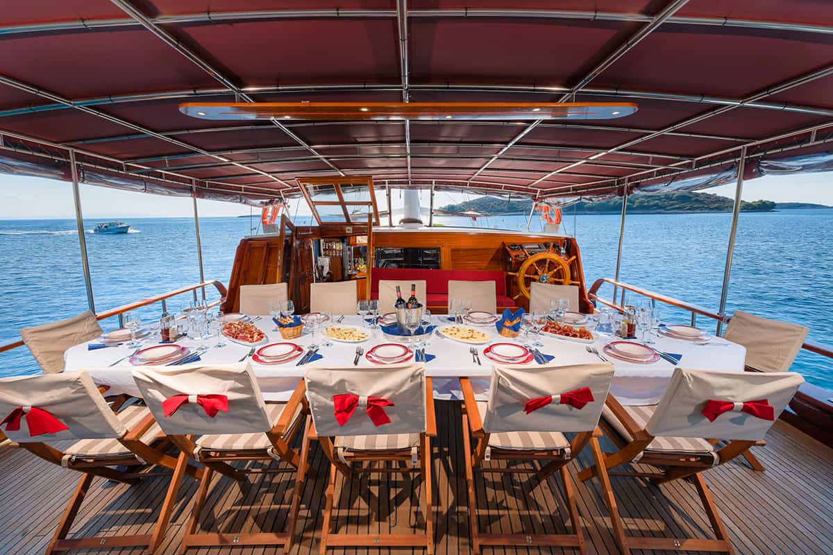 A long dining table set with plates, glasses, and food is arranged on a yacht deck, surrounded by wooden chairs with white covers and red bows, overlooking calm blue water and distant islands.