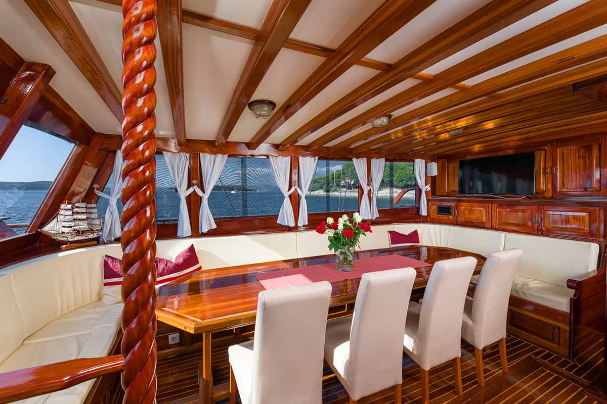The image shows a luxurious yacht interior with polished wood, a long table, white cushioned chairs, large windows with curtains, and a vase of red and white flowers on the table. Water and trees are visible outside.