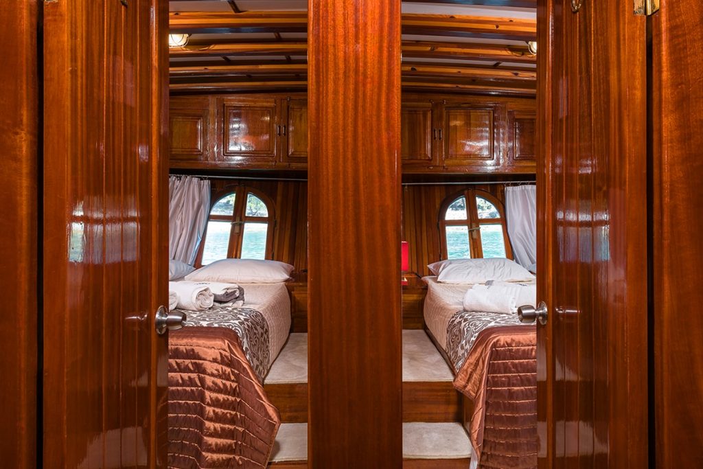 A view through double doors reveals a cozy boat cabin with two single beds, wooden walls, round windows with white curtains, and folded towels on the beds. Natural light streams in from the outside.