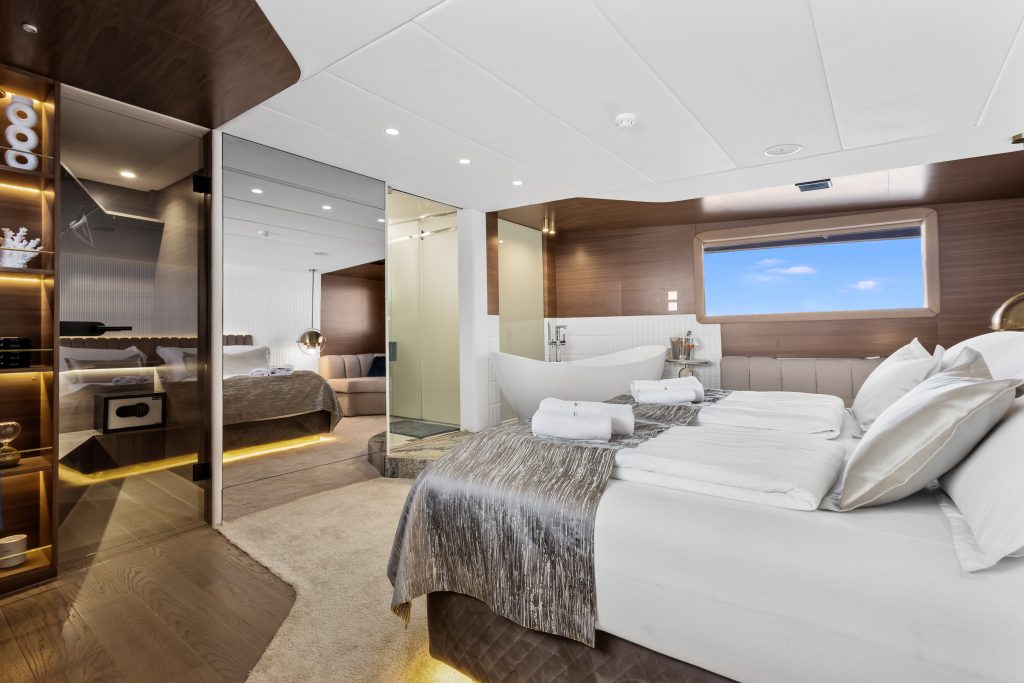A luxurious yacht bedroom suite with a large bed, elegant lighting, wood accents, a freestanding bathtub near a window, and modern decor featuring neutral tones and ocean views.