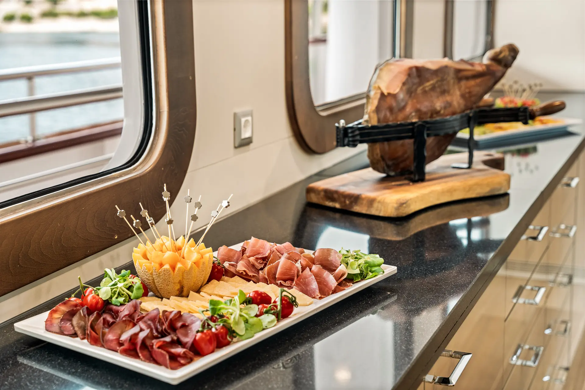 A platter of assorted cured meats, cheese, melon balls on skewers, cherry tomatoes, and greens sits on a counter next to a large leg of cured ham mounted on a stand, with windows and a view of water in the background.