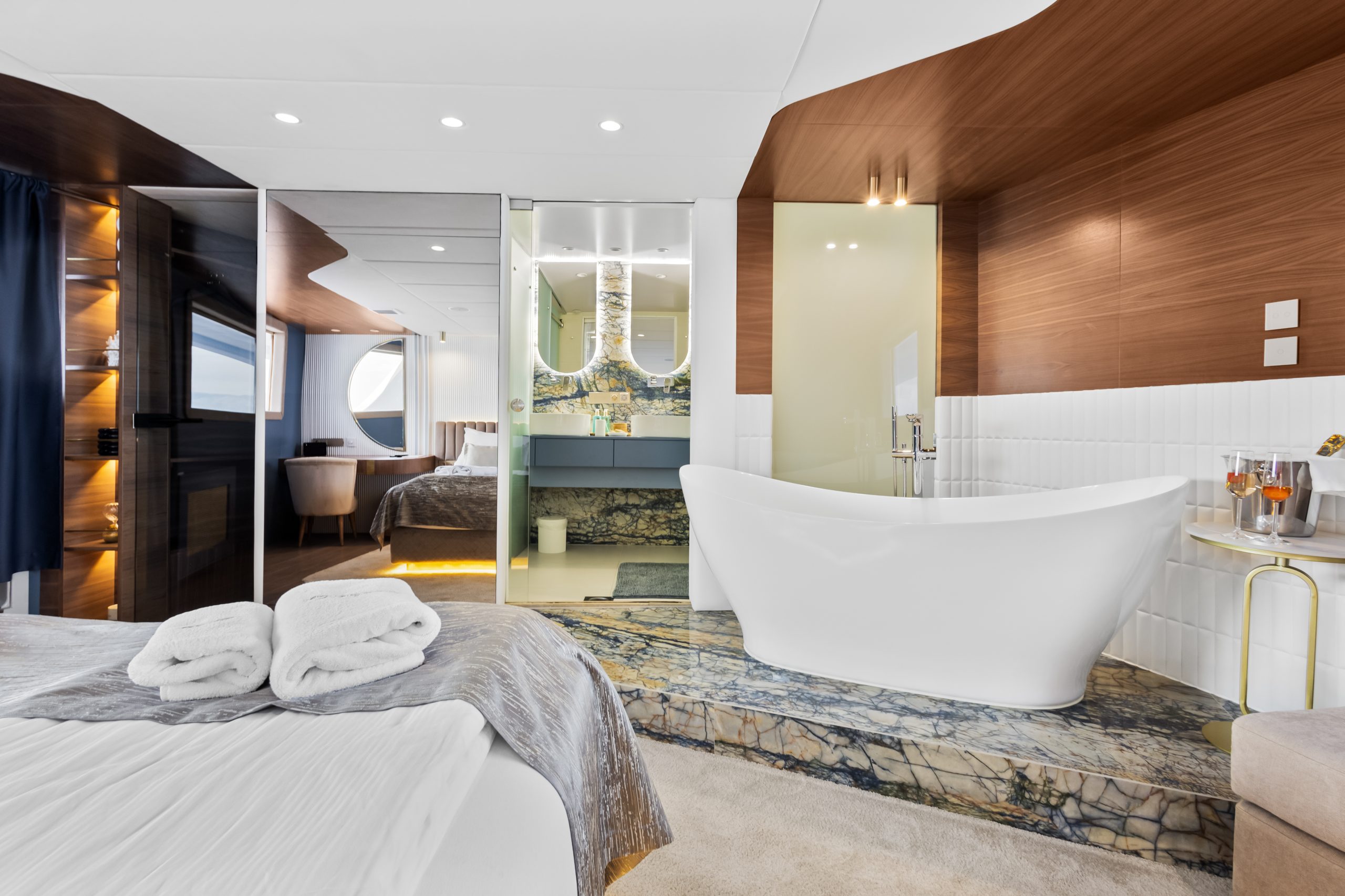 Modern hotel suite with a freestanding bathtub on marble flooring, bed with folded towels, sleek wood paneling, and a mirrored vanity area. A sitting area and large window are visible in the background.