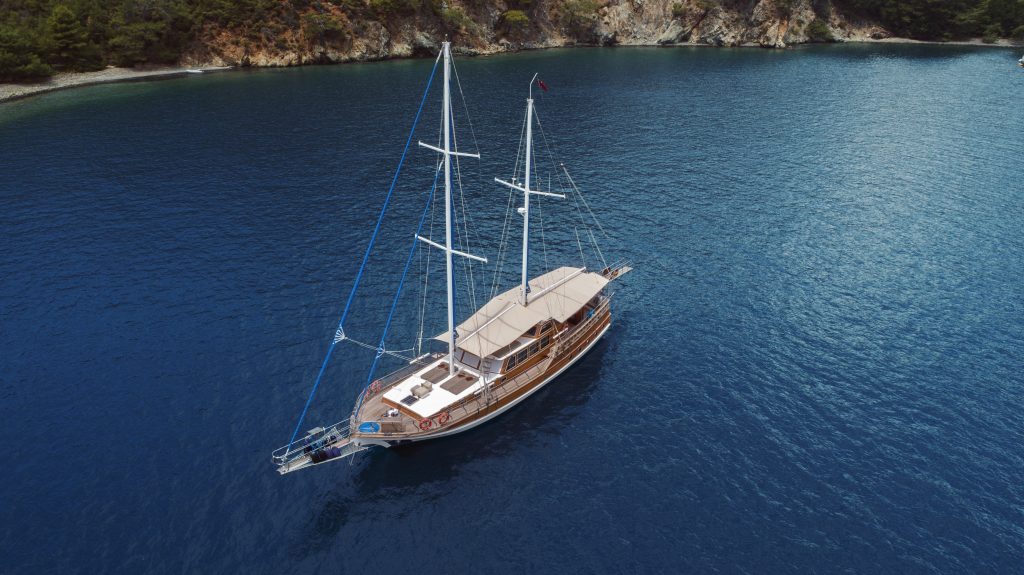 Aerial view of a wooden sailboat with two masts anchored in calm, deep blue water near a forested rocky coastline. The boat has a covered deck and white seating areas.