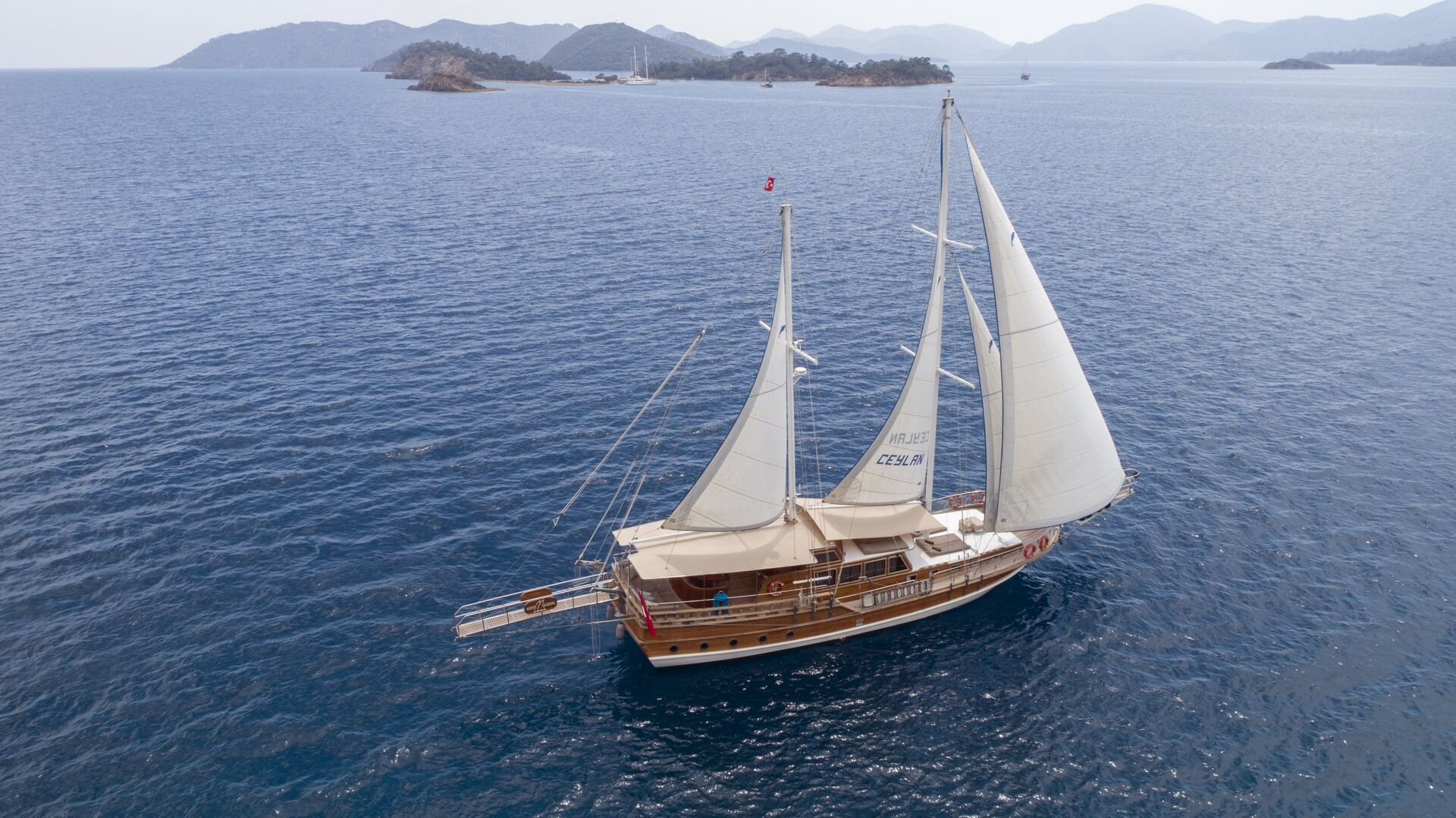 A wooden sailboat with white sails glides on calm blue sea waters, surrounded by small green islands and distant mountains under a light, hazy sky.