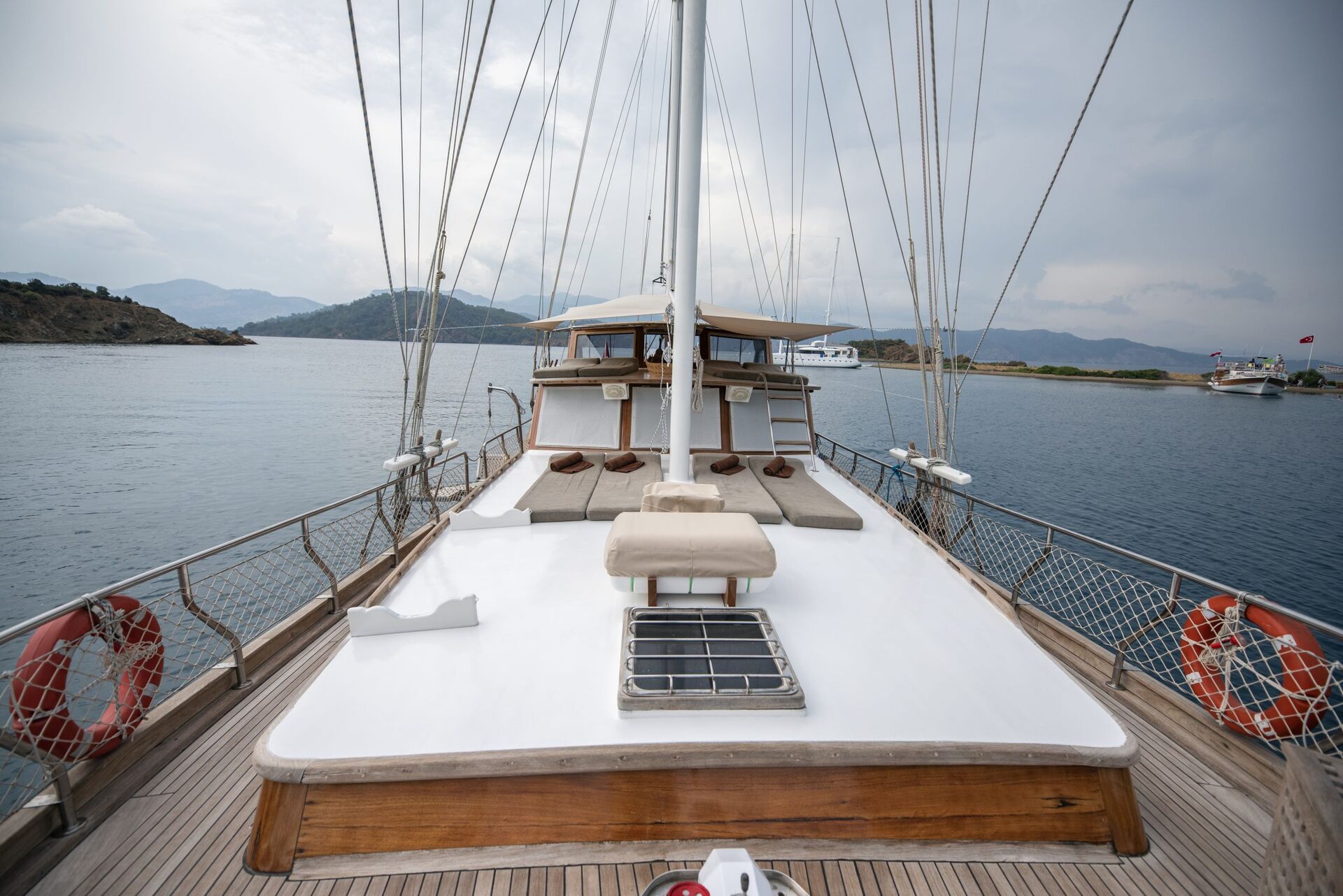 View from the deck of a sailboat with cushioned seating areas, looking toward the cabin; the boat is on calm water near islands under a cloudy sky.