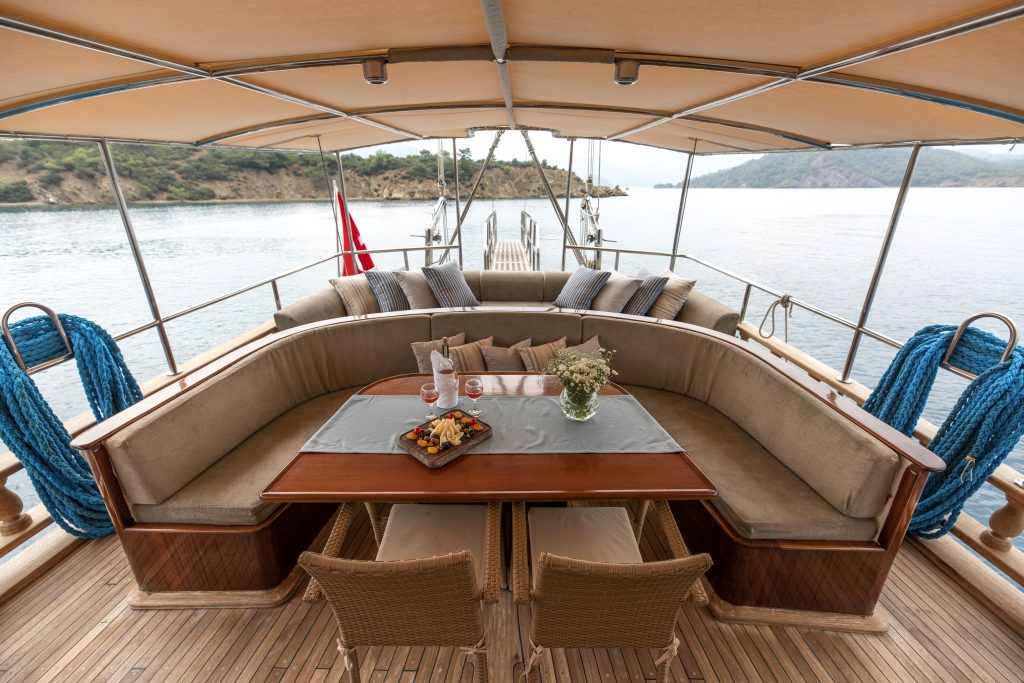 Una cubierta de yate con un gran sofá curvado, una mesa de madera para bebidas y aperitivos, dos sillas, cojines decorativos y cuerdas azules. El yate está anclado cerca de una costa tranquila con colinas al fondo.