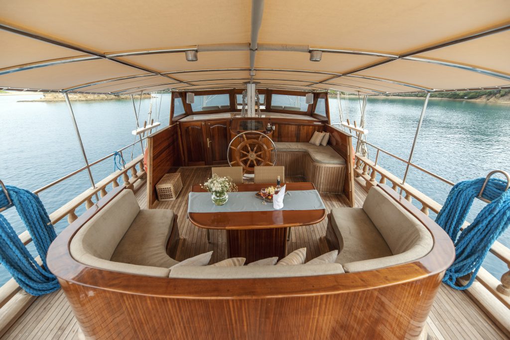 Una lujosa cubierta de yate con asientos de madera curvada, una mesa con un jarrón de flores y vasos, un volante de madera y un toldo cubierto, rodeada de aguas tranquilas. A ambos lados hay cuerdas azules cuidadosamente enrolladas.