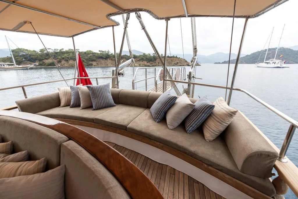 A curved beige sofa with striped and solid pillows sits on the deck of a yacht, overlooking calm water and distant green hills. Other boats and a red flag are visible in the background under an overcast sky.