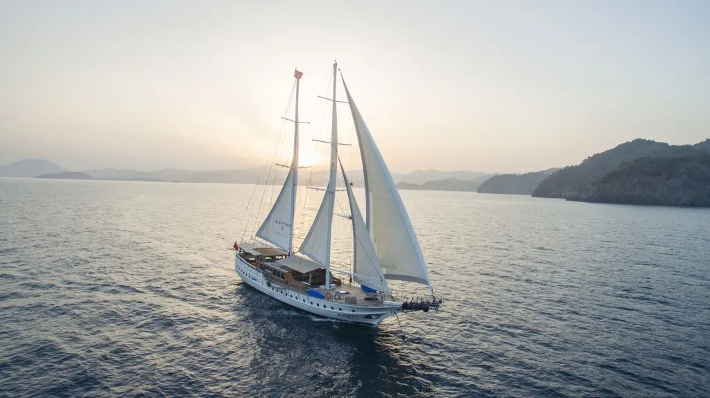 A white sailboat with two large sails glides across calm blue water at sunset, with distant mountains and a hazy sky in the background.