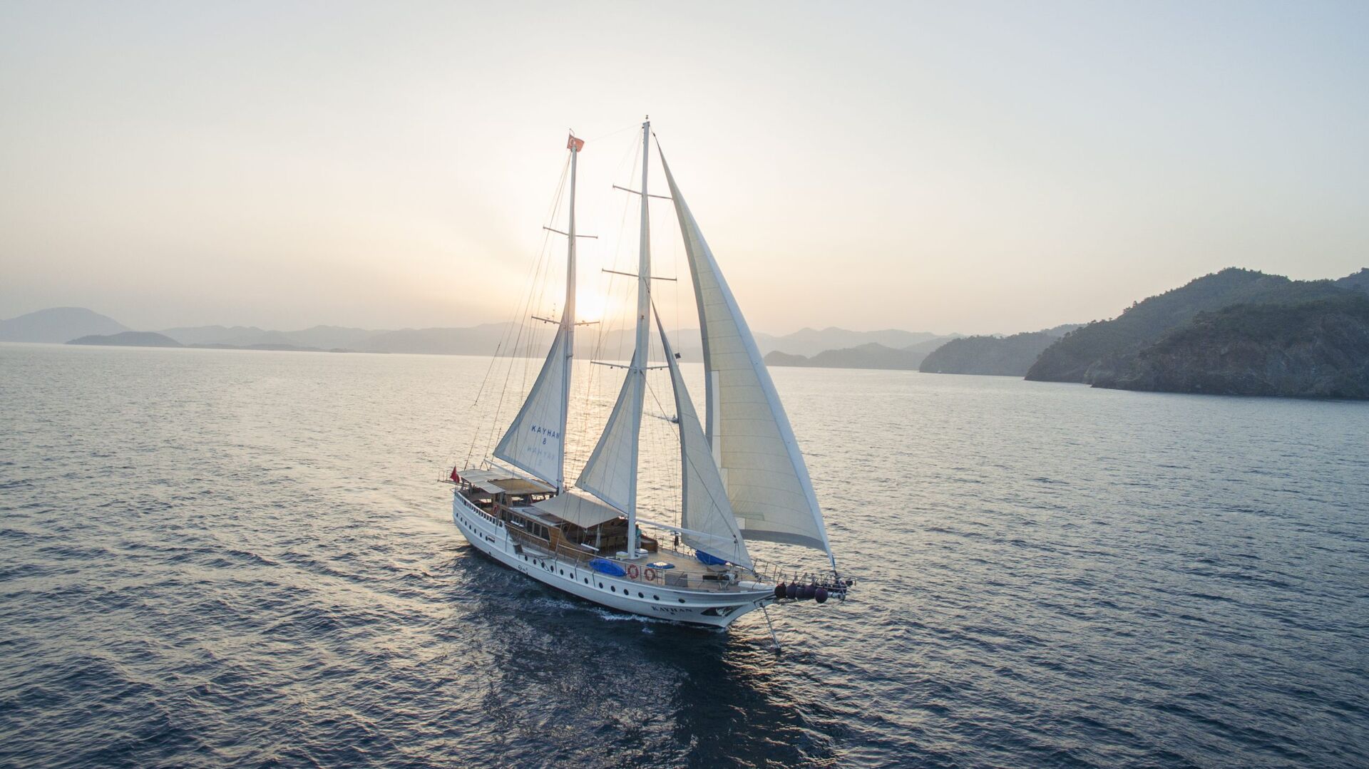 A white sailboat with two large sails glides across calm blue water at sunset, with distant mountains and a hazy sky in the background.