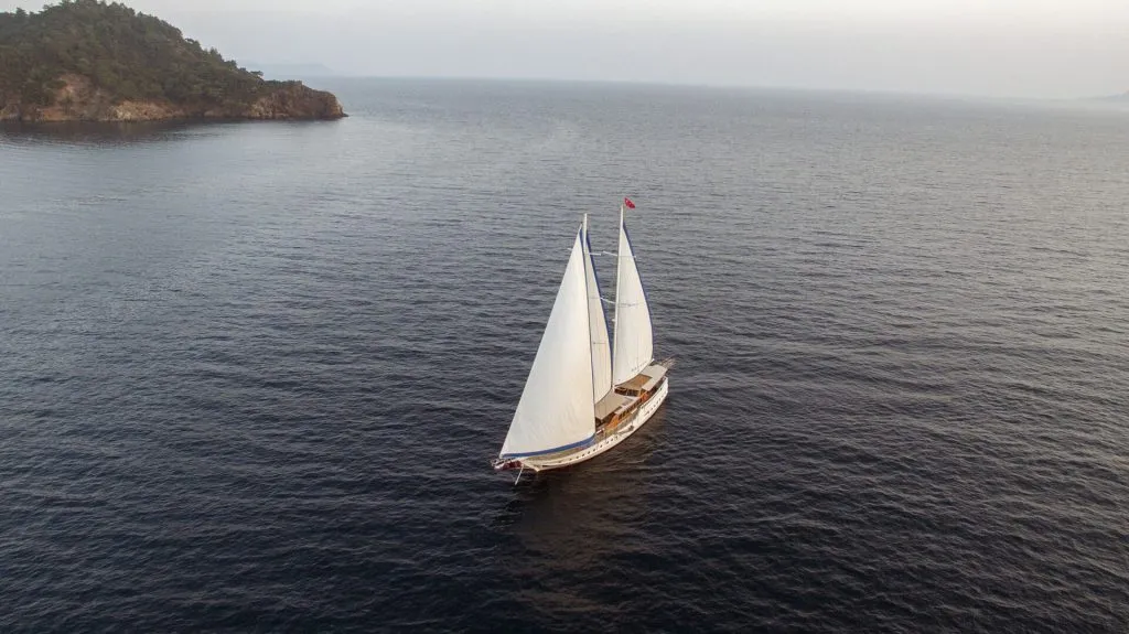 Aerial view of a sailboat with white sails gliding on calm, dark blue water near a tree-covered coastline under a cloudy sky.