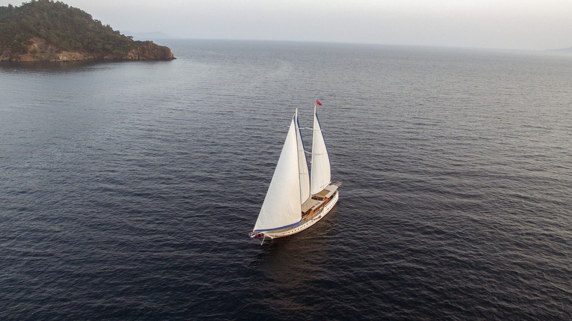 Aerial view of a sailboat with white sails gliding on calm, dark blue water near a tree-covered coastline under a cloudy sky.