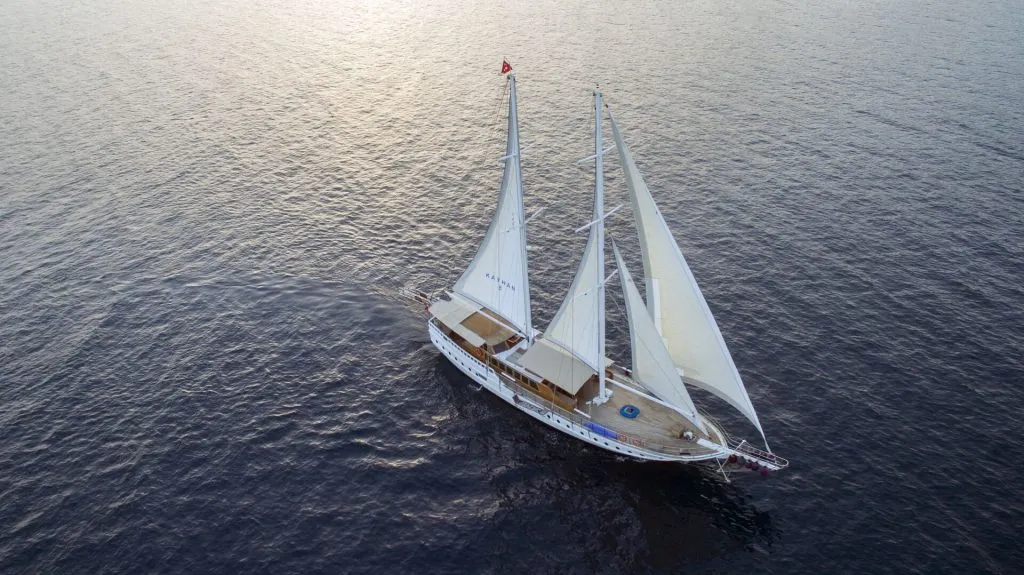 Aerial view of a white sailing yacht with three tall sails gliding smoothly over calm, dark blue water, leaving a gentle wake behind on a bright, serene day.