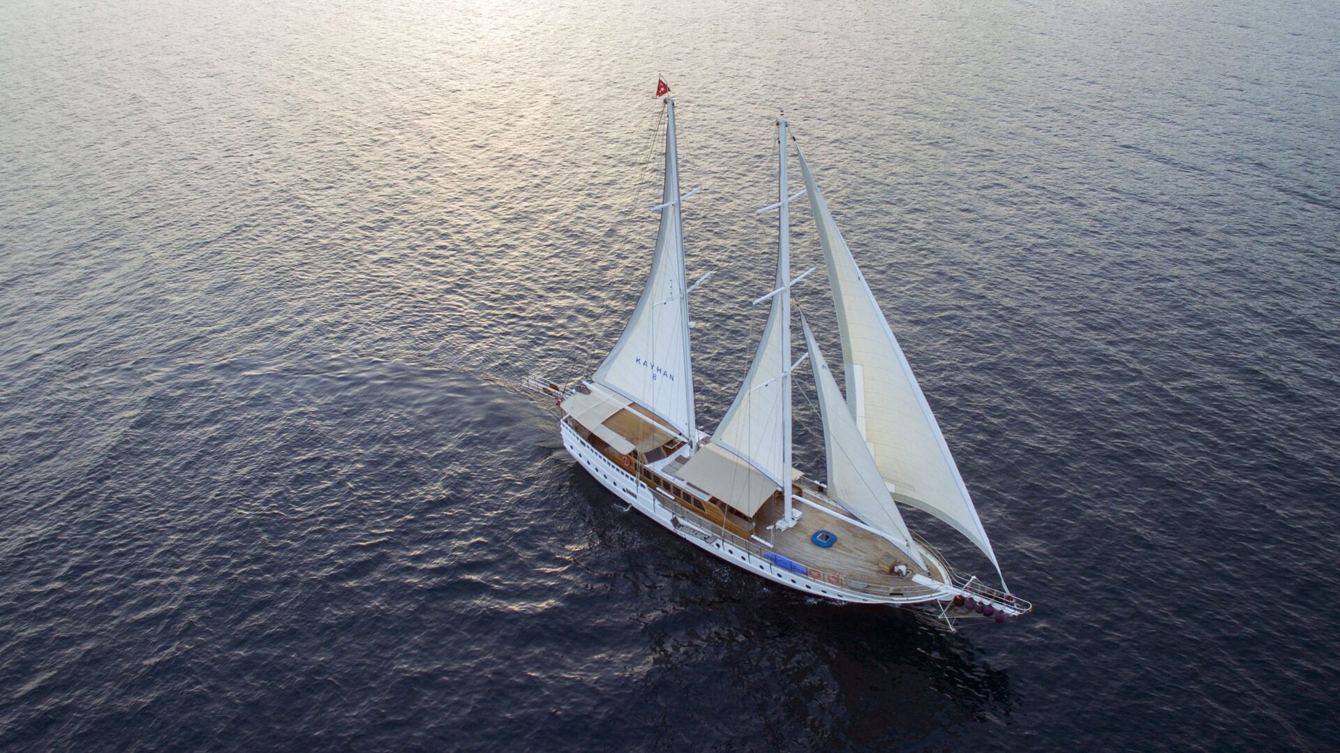 Aerial view of a white sailing yacht with three tall sails gliding smoothly over calm, dark blue water, leaving a gentle wake behind on a bright, serene day.