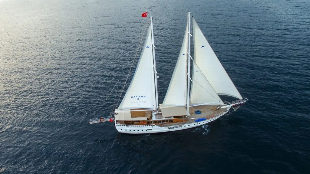 A white sailing yacht with two sails is moving across calm, dark blue water. The yacht has a Turkish flag at the stern and another on the main mast. The deck is wooden, and the yacht is seen from above.