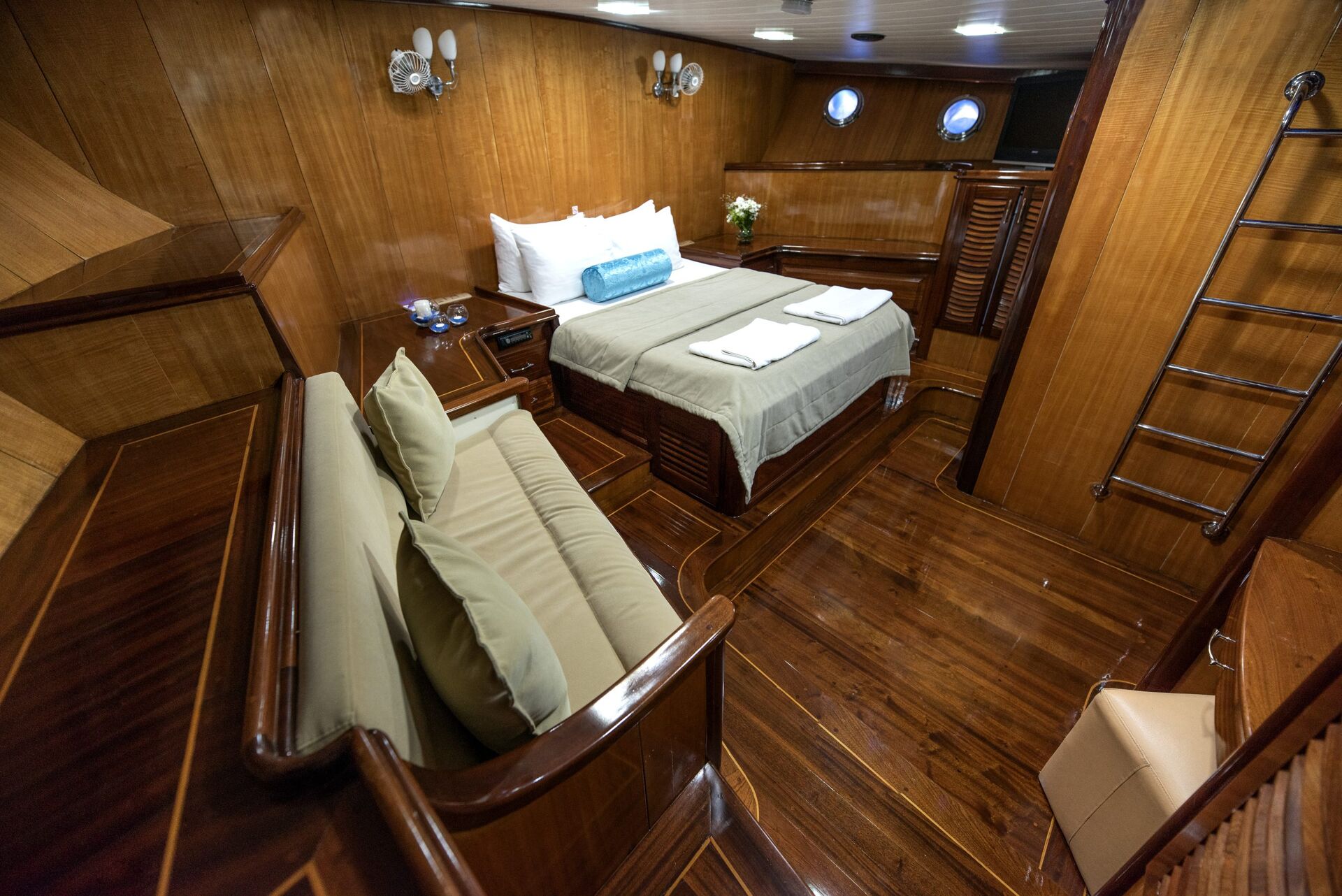 Luxurious ship cabin with polished wood walls and floor, a double bed with towels, a sofa, nightstands, round windows, and a metal ladder on the right wall. A vase of flowers sits on a side table.