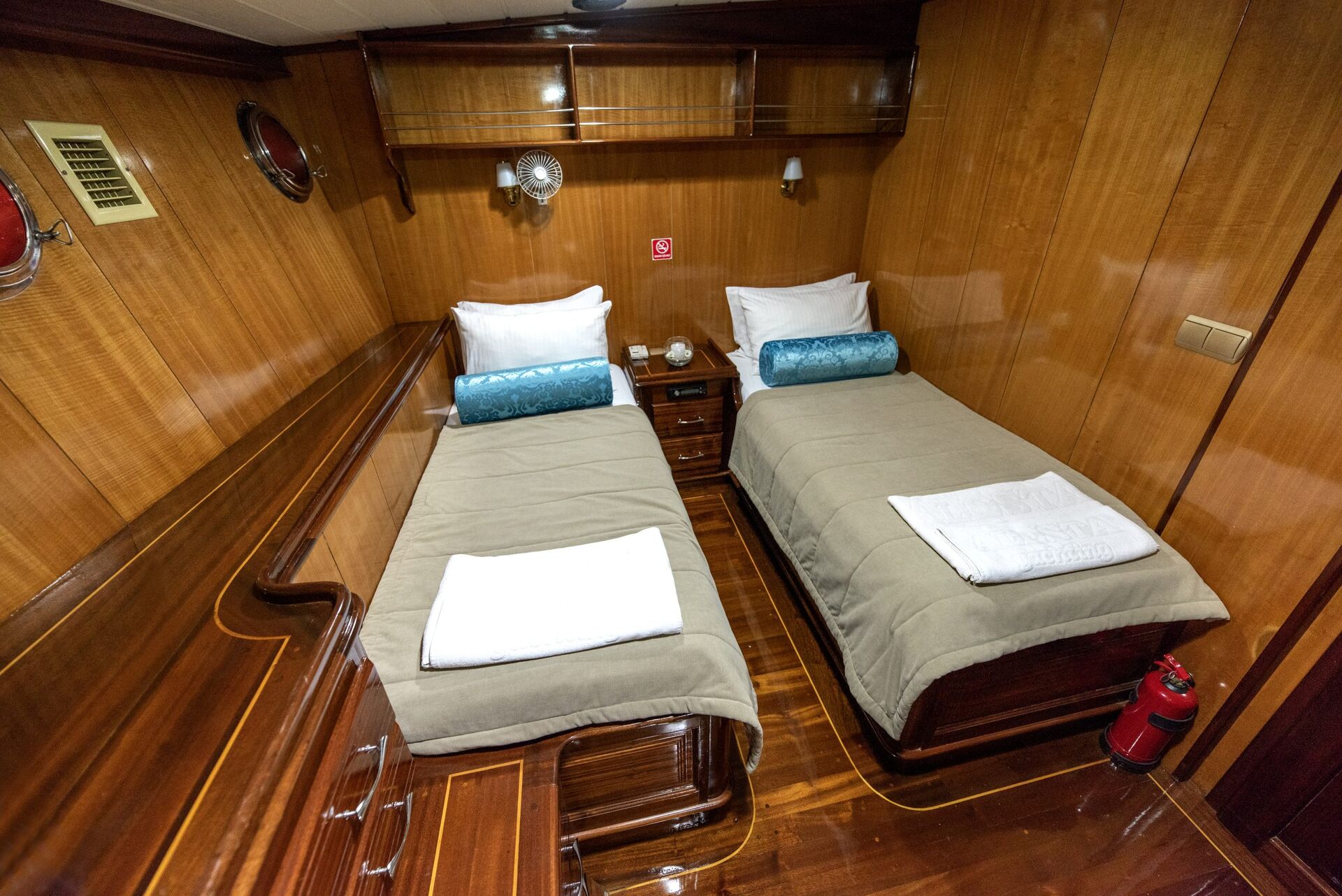 A cozy wooden cabin with two single beds, each with beige bedding, white pillows, and blue cylindrical cushions. A nightstand separates the beds, and a fire extinguisher is on the floor by the right bed.