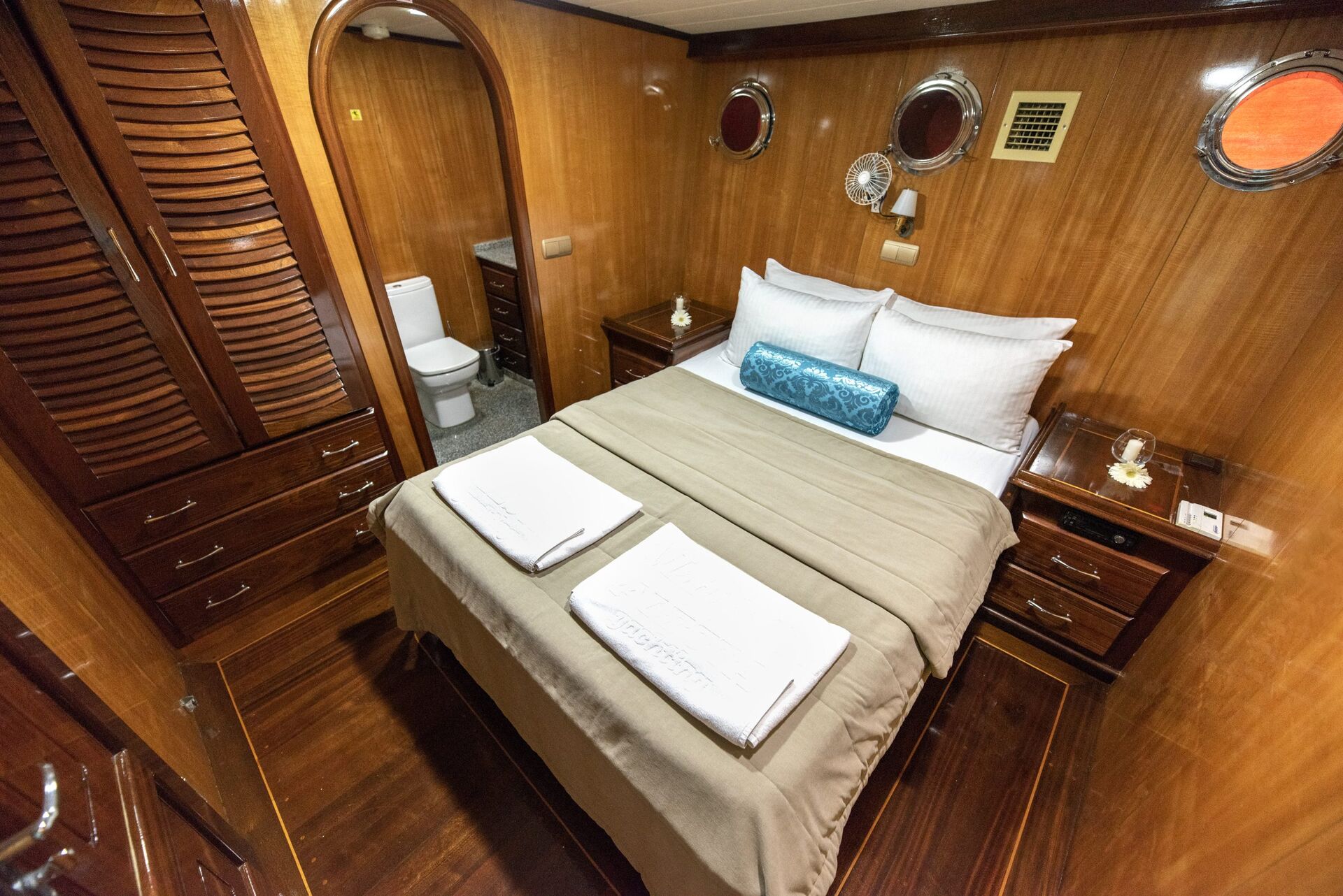 A cozy yacht cabin with wood-paneled walls, a double bed neatly made with white linens and two folded towels, bedside tables with lamps, and a visible en-suite bathroom with a toilet.