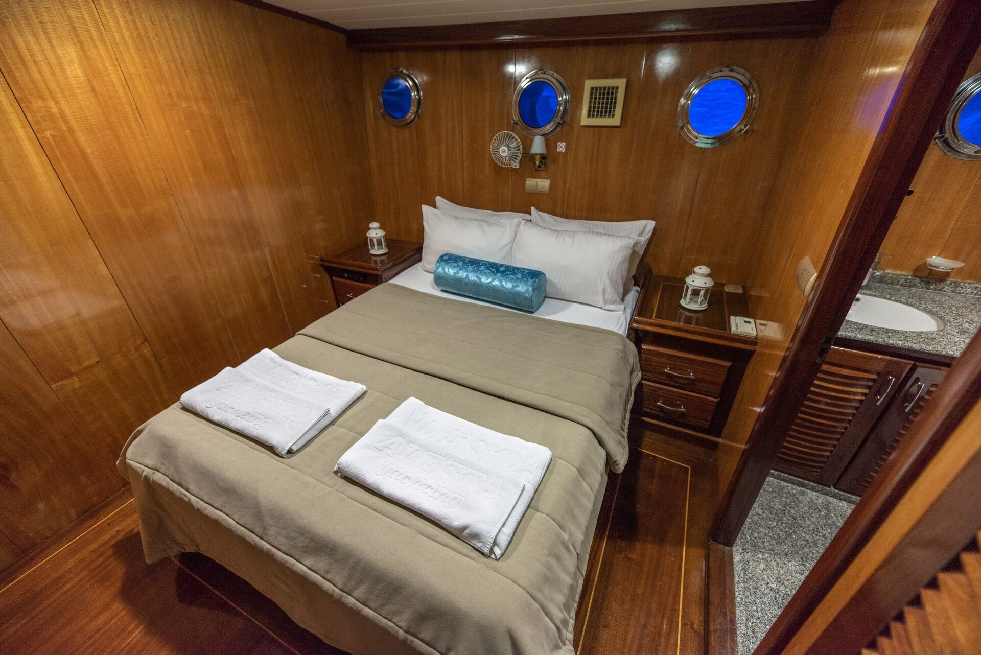 A cozy cabin bedroom with wood-paneled walls, a double bed neatly made with beige linens and two towels, small round windows, bedside tables with lamps, and a countertop with a sink to the right.
