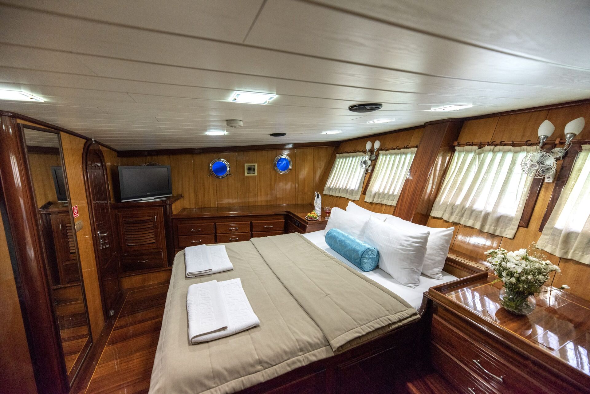 A cozy, wood-paneled cabin bedroom with a large bed, white and beige bedding, rolled blue pillow, two folded towels, a TV, drawers, portholes, curtains, lamps, and a vase of flowers on a nightstand.