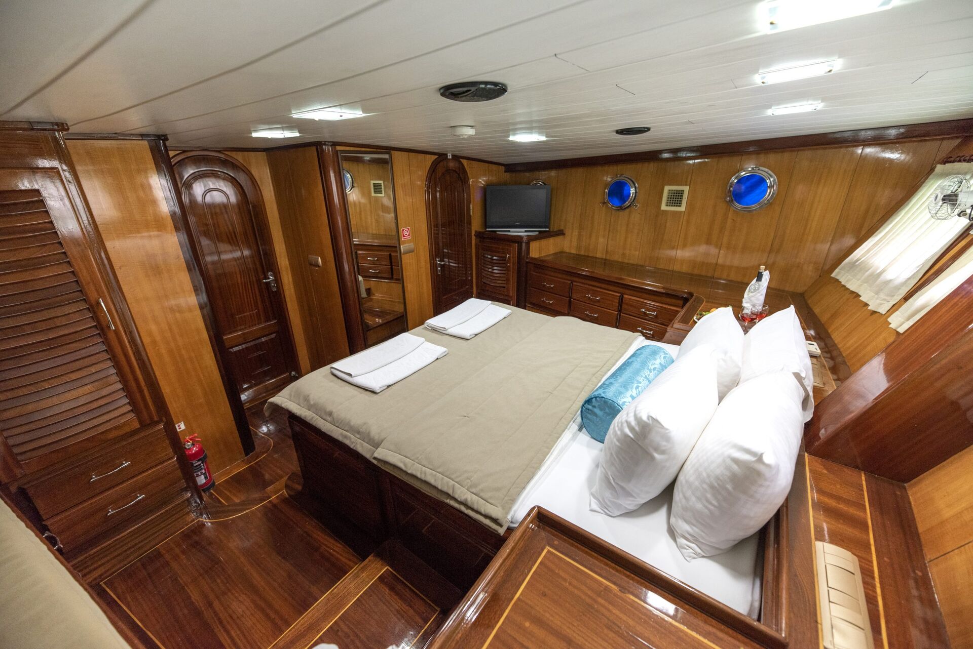 A yacht cabin with polished wooden walls and floor, a neatly made double bed with white pillows and beige bedding, a TV, cabinets, porthole windows, and natural light coming through a curtained window.