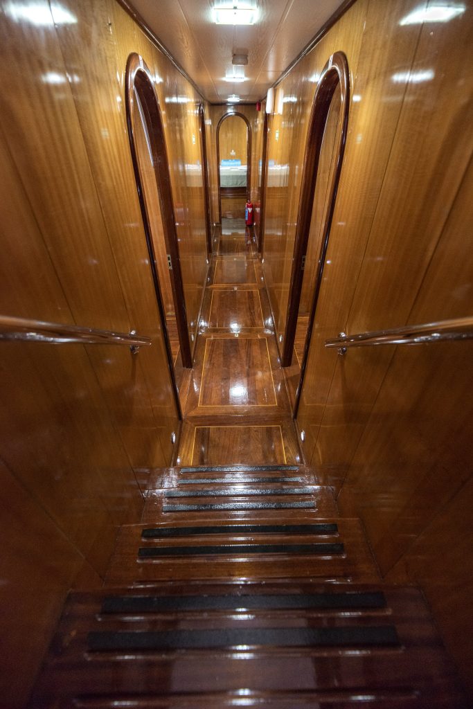 View down a narrow, polished wooden hallway with steps leading to doorways on either side and a small sink visible at the end. Bright lights reflect off the shiny wood surfaces.