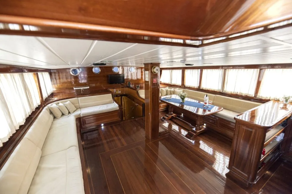Luxurious yacht interior with polished wooden floors, cream-colored sofas lining the walls, a dining table with chairs, and large windows with curtains letting in natural light.