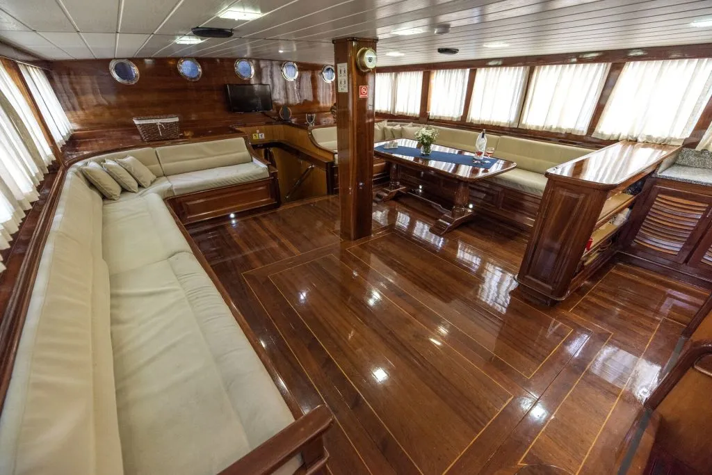 Spacious yacht interior with polished wooden floors, cream-colored sofas, a dining area with cushioned benches, large windows with curtains, and a small table with a plant centerpiece.