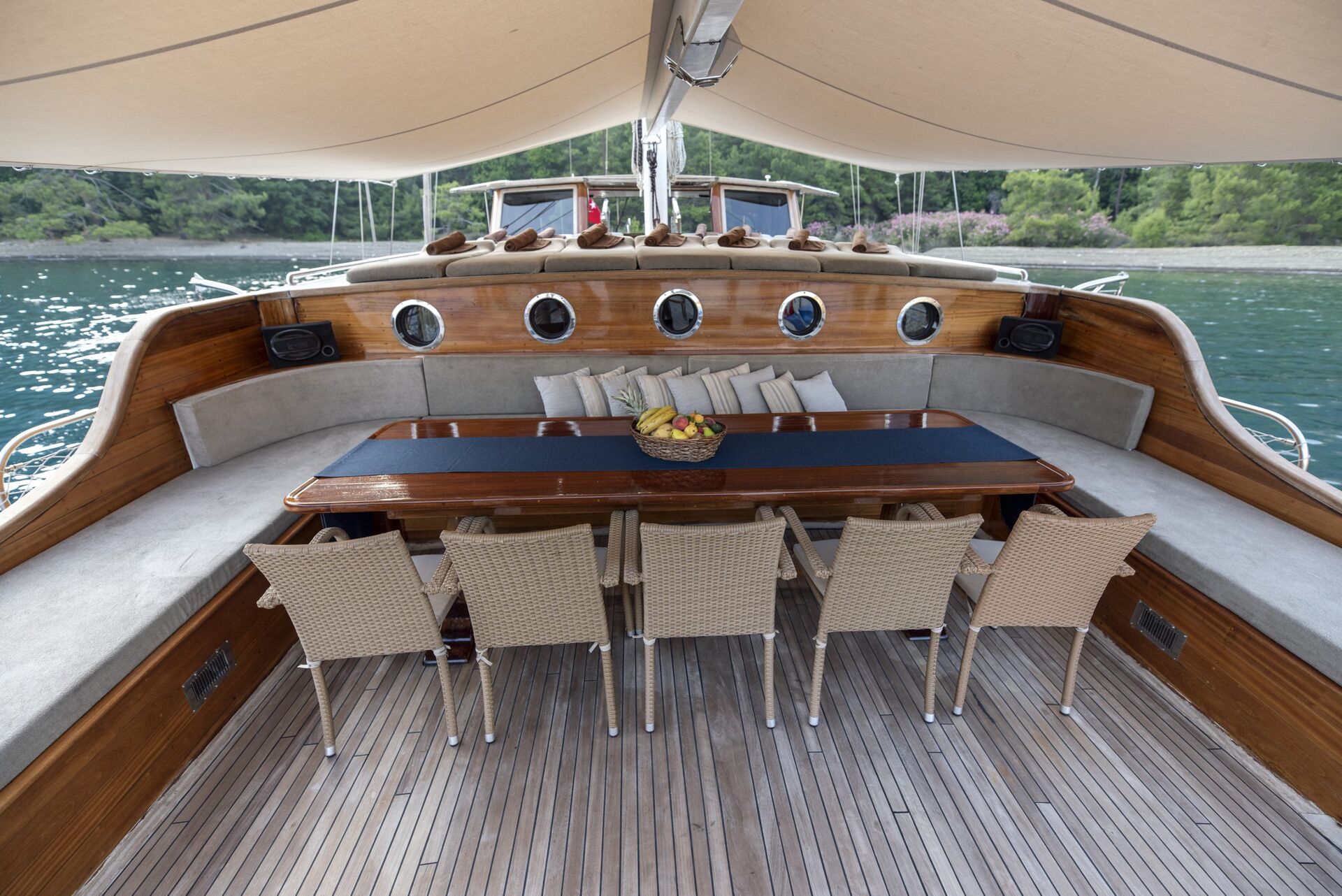 A luxurious yacht deck features a curved cushioned bench, eight wicker chairs, a long wooden table with a fruit basket, and a tan canopy, with water and trees visible in the background.
