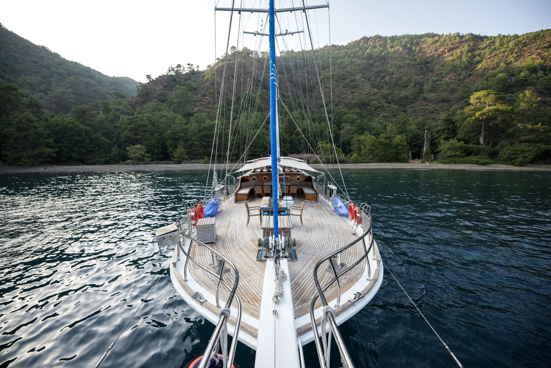 A view from the bow of a sailboat anchored near a wooded shoreline, with calm water and tree-covered hills in the background under a clear sky.