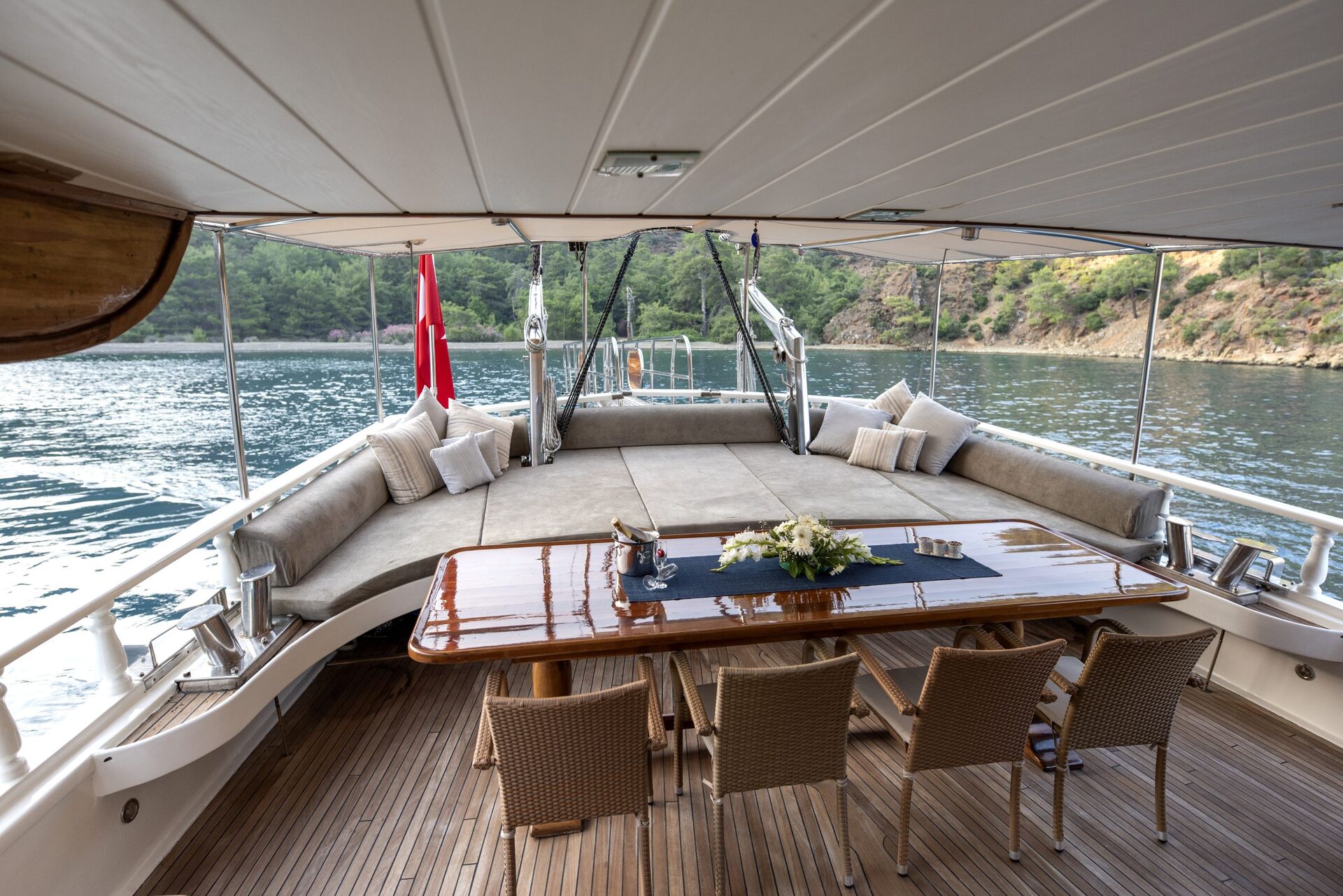 Outdoor dining area on a yacht with a polished wooden table, wicker chairs, cushioned lounge beds, and scenic views of calm water and green hills in the background.