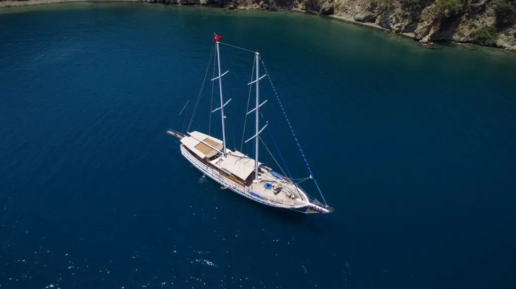 Aerial view of a white sailing yacht with three masts anchored in calm, deep blue water near a rocky, green shoreline. A red flag is visible at the stern.