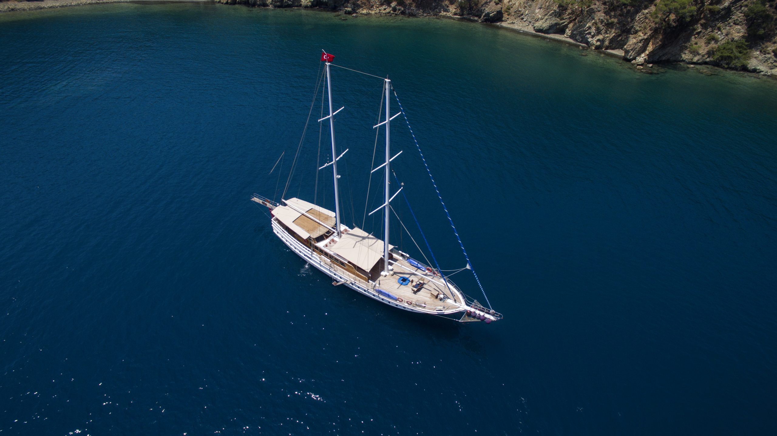 Aerial view of a white sailing yacht with three masts anchored in calm, deep blue water near a rocky, green shoreline. A red flag is visible at the stern.