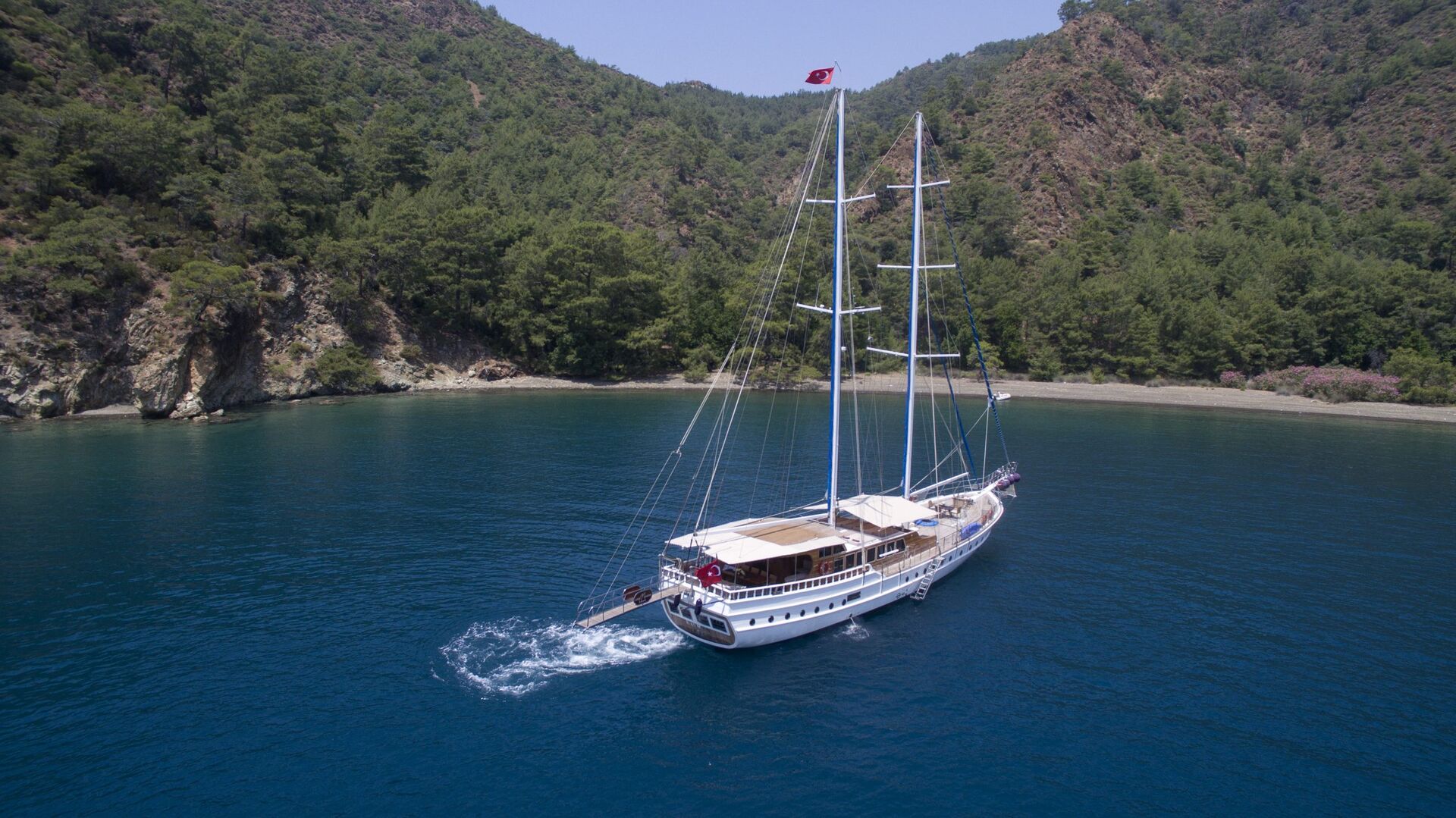 A large white sailboat with multiple masts sails on calm blue water near a forested, mountainous shoreline under a clear sky.