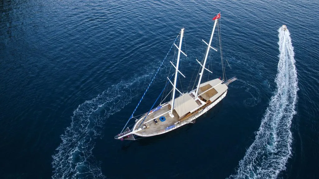 Aerial view of a sailboat with three masts sailing on deep blue water, while a small motorboat creates a curved wake nearby.