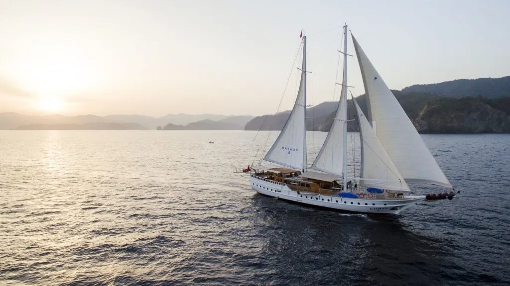 A white sailing yacht with large sails glides across calm sea waters at sunset, with mountains and a hazy sky in the background.