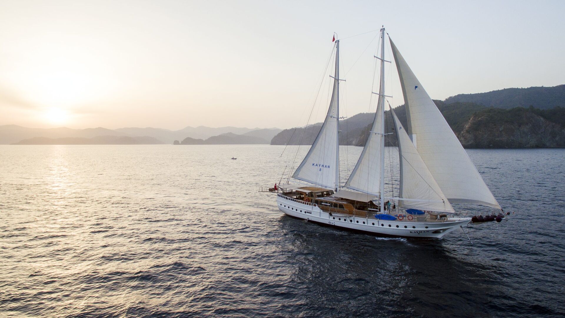 A white sailing yacht with large sails glides across calm sea waters at sunset, with mountains and a hazy sky in the background.