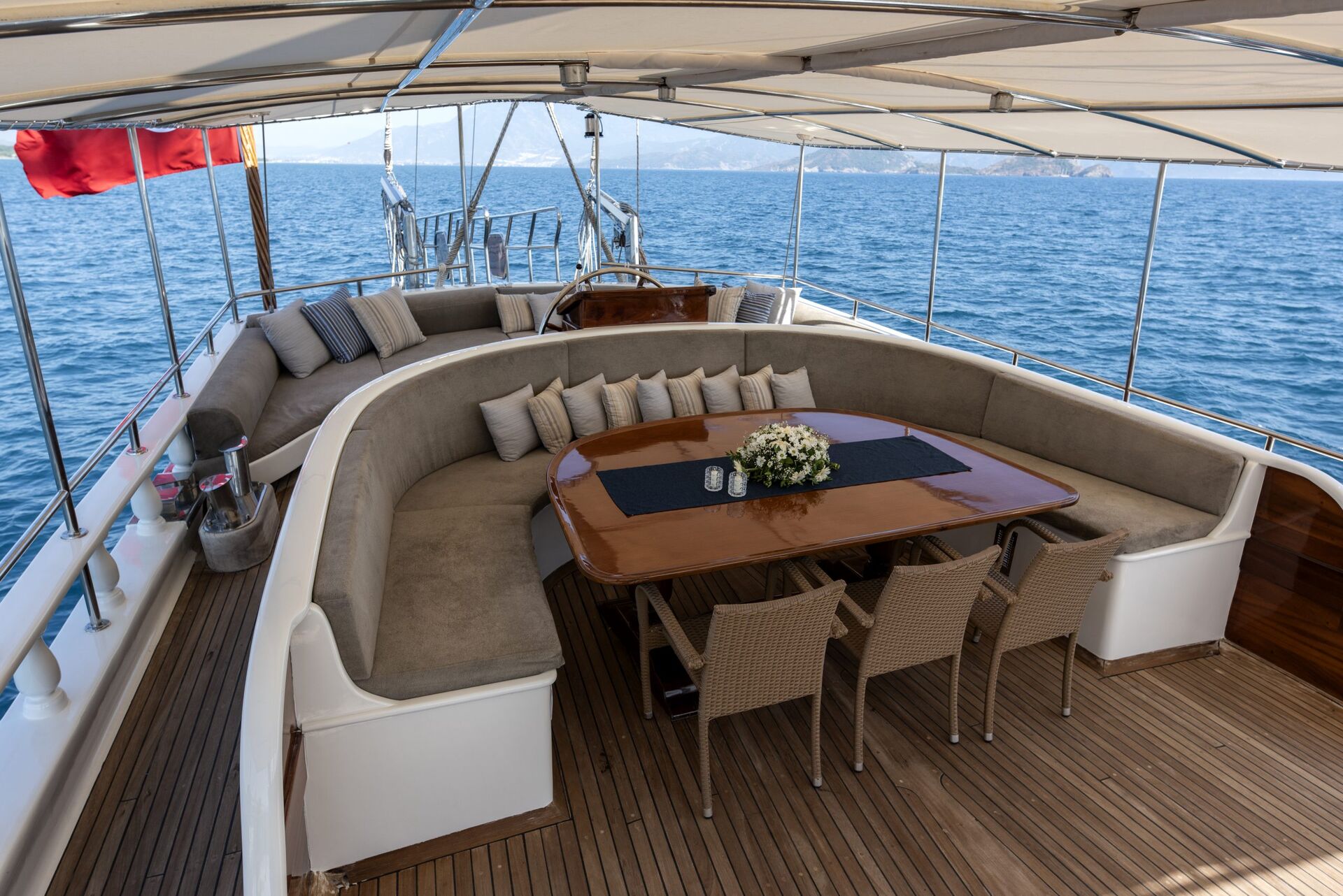 A luxurious yacht deck with a curved beige sofa, a wooden dining table set with six chairs, a floral centerpiece, and a shaded canopy, overlooking calm blue sea under a clear sky.