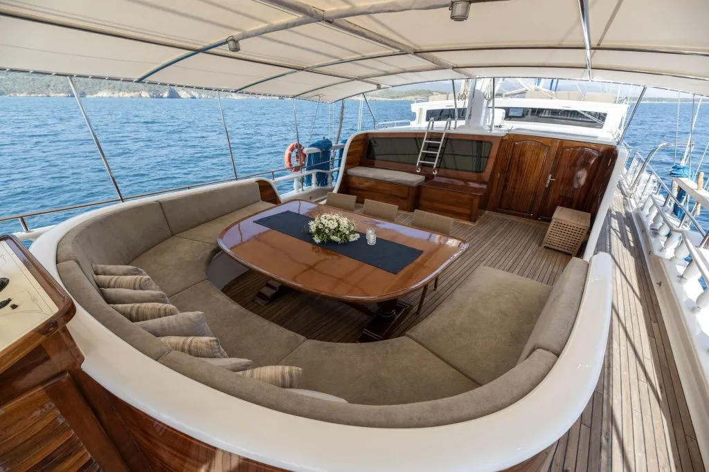 A luxury yacht deck with a curved beige sofa around a polished wooden table, decorated with flowers. The yacht is on calm blue water, with land visible in the distance under a canopy.
