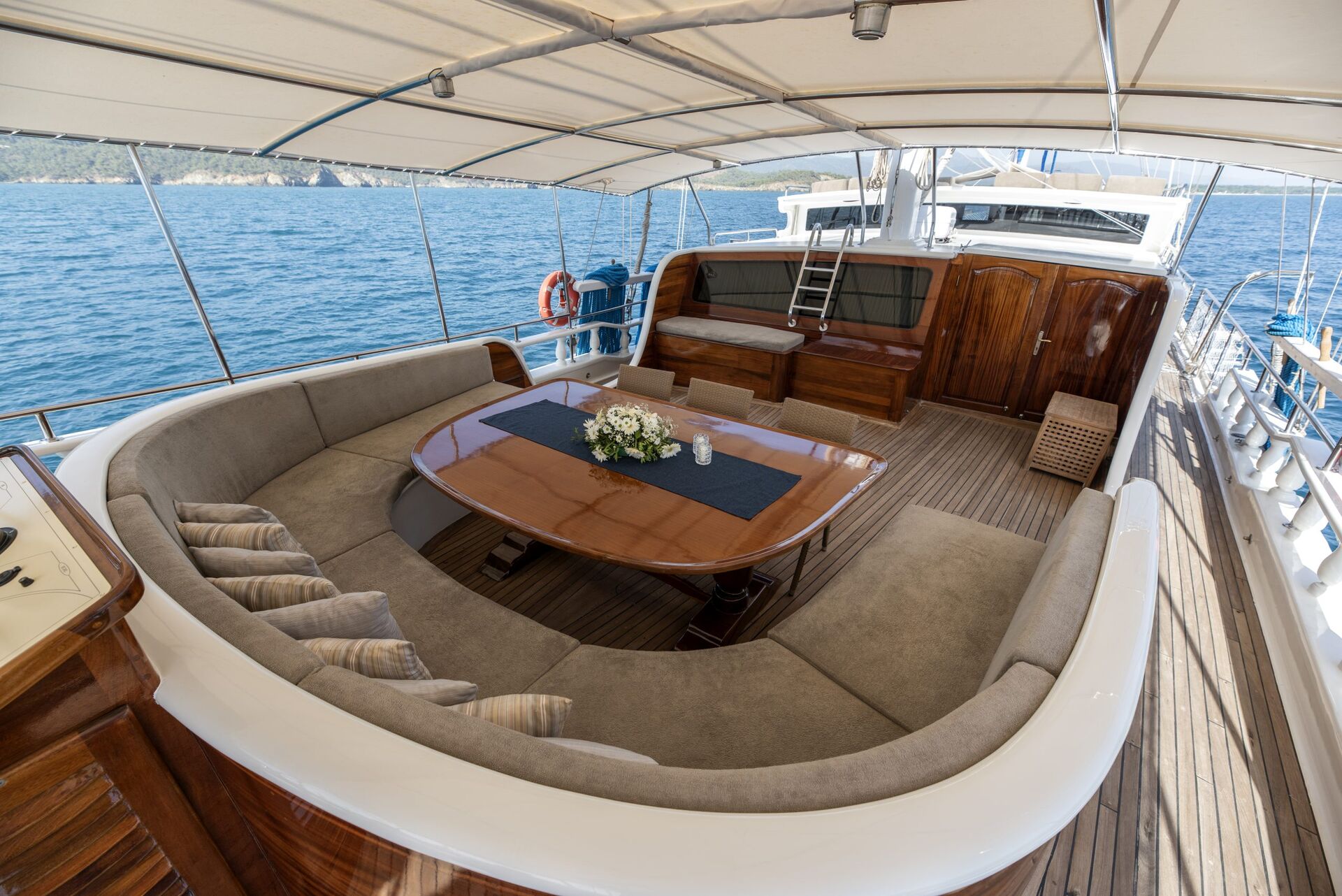A luxury yacht deck with a curved beige sofa around a polished wooden table, decorated with flowers. The yacht is on calm blue water, with land visible in the distance under a canopy.