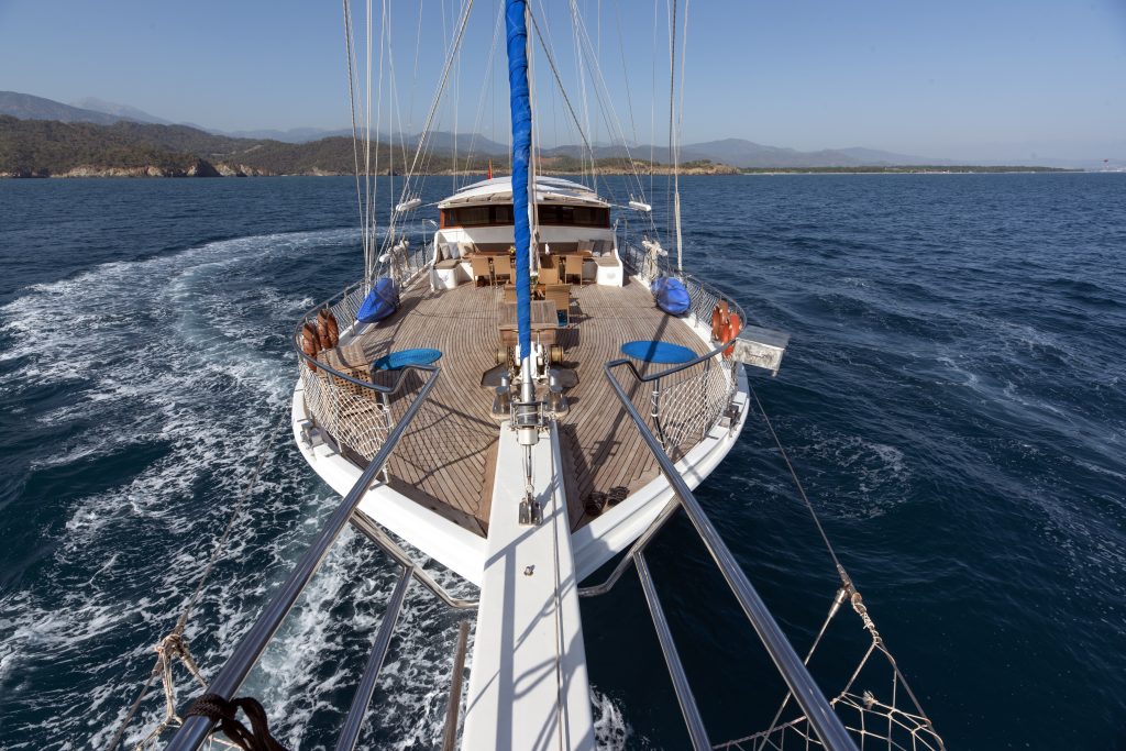 View from the bow of a yacht moving through blue ocean water, with a spacious wooden deck, seating areas, and mountains visible in the distance under a clear sky.