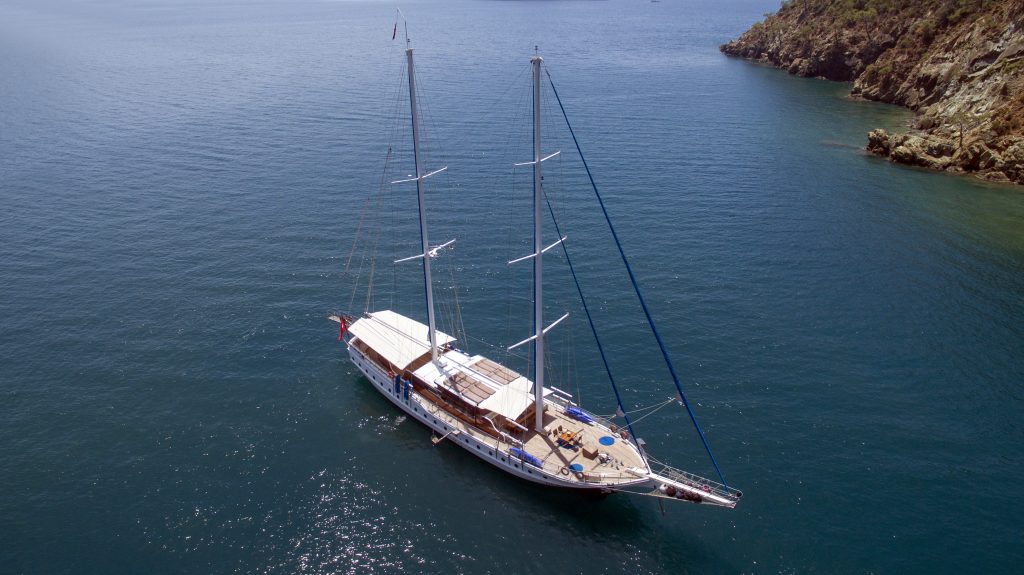 A white sailboat with two tall masts floats on calm blue water near a rocky shoreline under a clear sky.