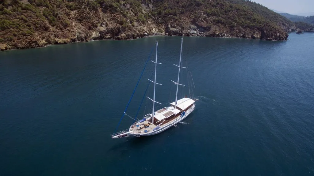 A white sailboat with three masts floats on calm blue water near a rocky, tree-covered shoreline under a clear sky.
