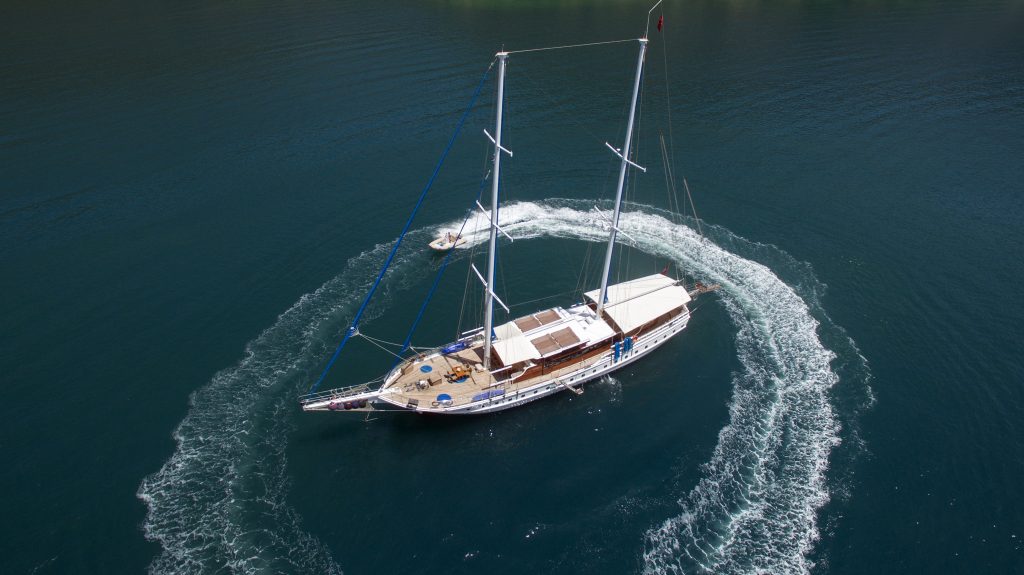 Aerial view of a large sailboat anchored in calm blue water, with a small motorboat creating circular waves around it.