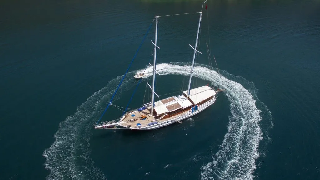 Aerial view of a large sailboat anchored in calm blue water, with a small motorboat creating circular waves around it.