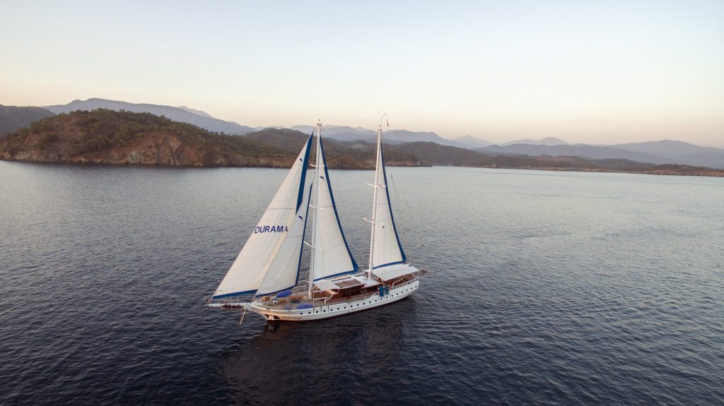 A white sailboat with two large sails glides across calm blue water near a rugged coastline, with hilly, tree-covered islands and mountains in the background under a clear sky at sunset.