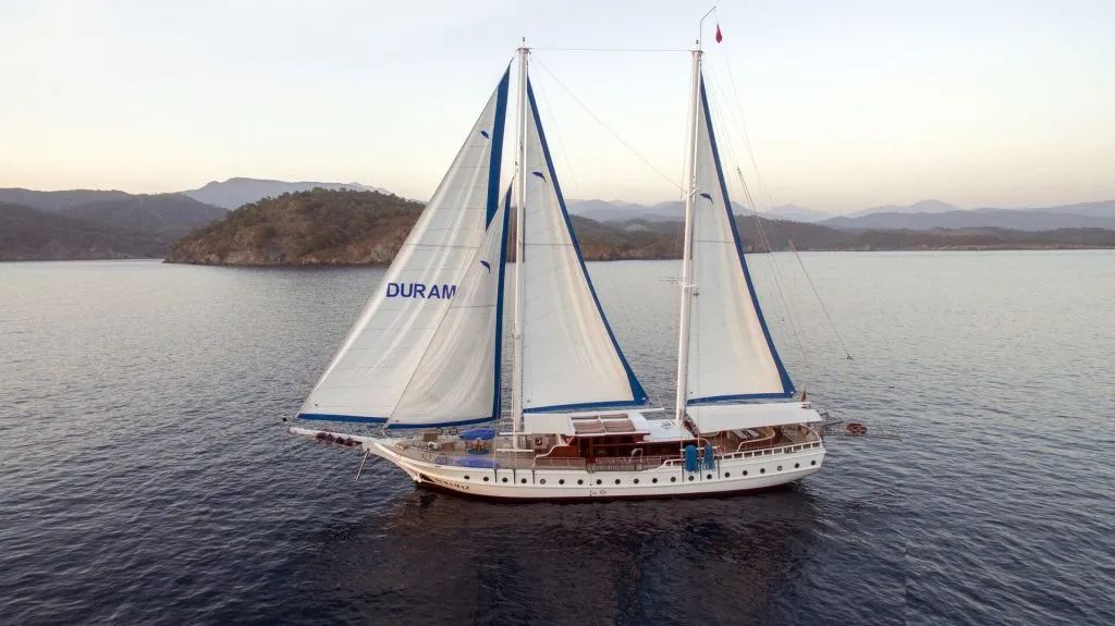 A white sailboat with two tall masts and blue trim sails on calm water near a forested coastline, with mountains visible in the background. The word DURAM appears on one of the sails.