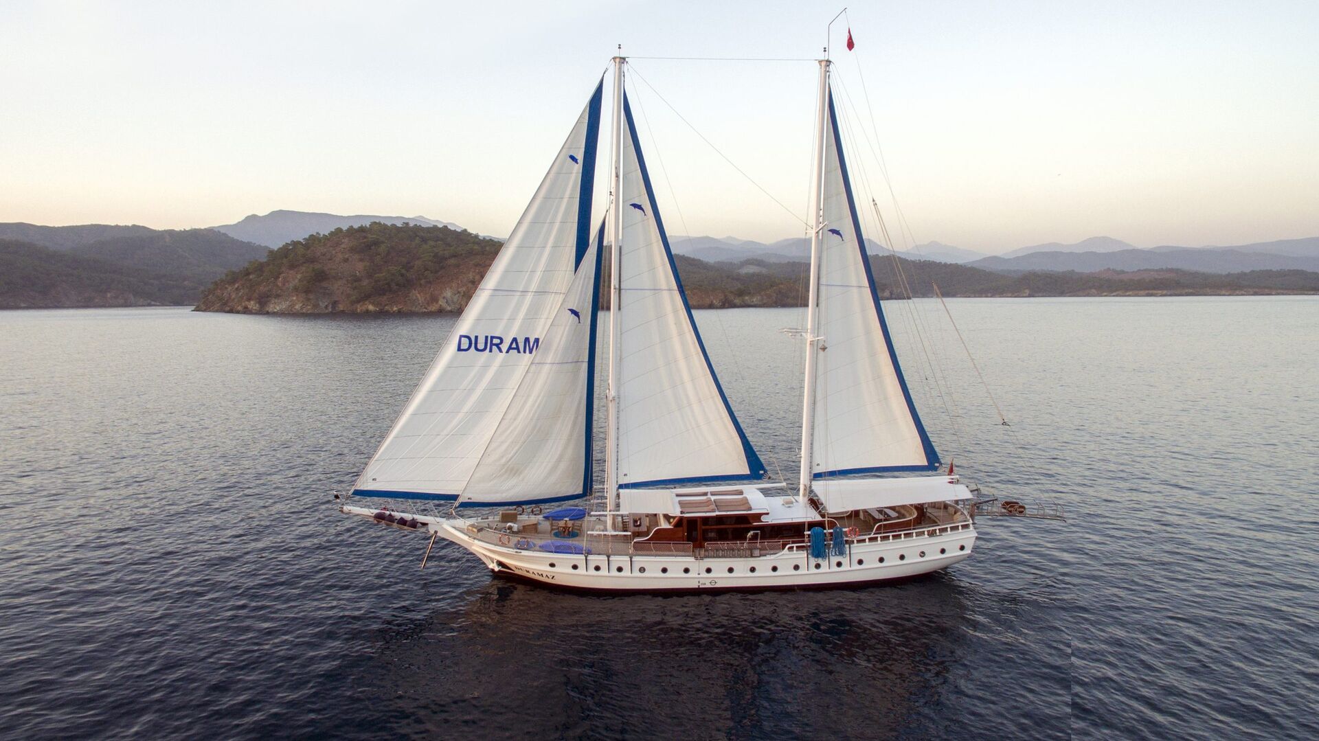 A white sailboat with two tall masts and blue trim sails on calm water near a forested coastline, with mountains visible in the background. The word DURAM appears on one of the sails.