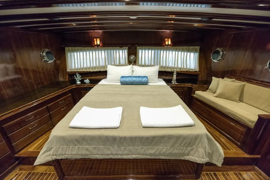 A cozy, wood-paneled cabin bedroom with a double bed, beige bedding, two white towels, a blue bolster pillow, side lamps, porthole windows, curtains, and built-in drawers with a cushioned bench.