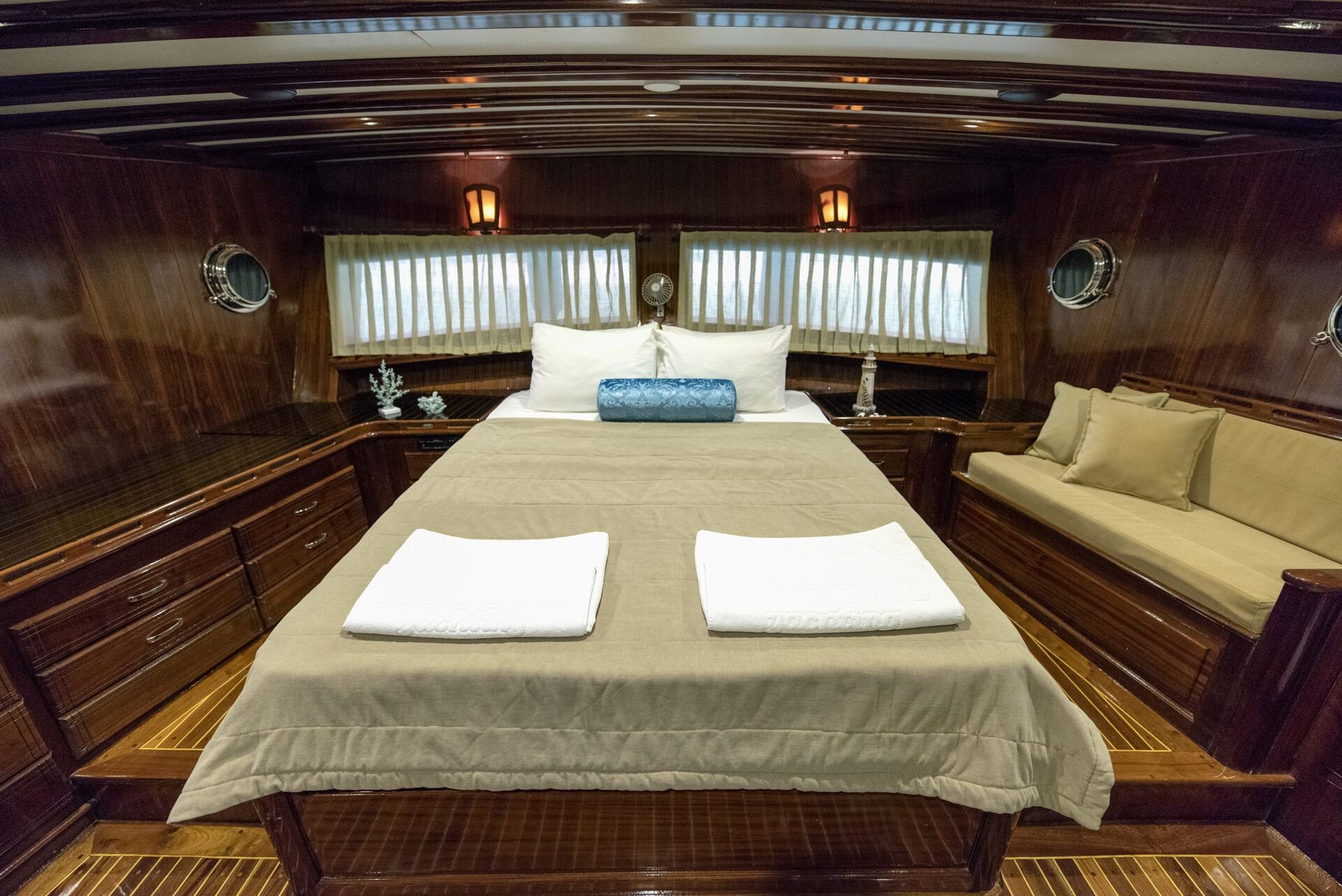 A cozy, wood-paneled cabin bedroom with a double bed, beige bedding, two white towels, a blue bolster pillow, side lamps, porthole windows, curtains, and built-in drawers with a cushioned bench.